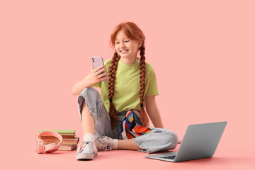 Cute ginger girl with modern laptop, mobile phone and books sitting on pink background