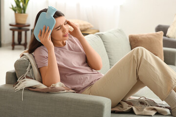 Young woman warming her head with hot water bottle on sofa in living room