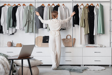 Young woman choosing clothes from wardrobe in light bedroom with cozy bed and laptop on bedside...