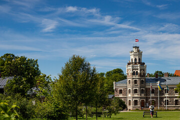Scenic summer view of a historic stone castle in Latvia with national flags, surrounded by trees and a clear blue sky.