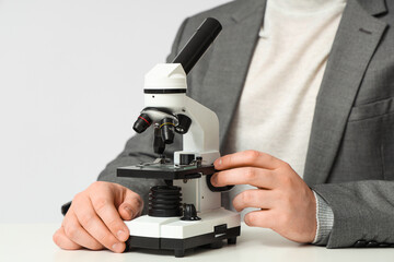 Young male teacher with microscope sitting at table on grey background, closeup