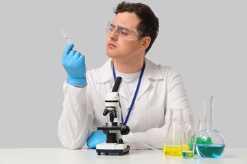 Thoughtful young male scientist with microscope, glass slide and chemical flasks sitting at table...