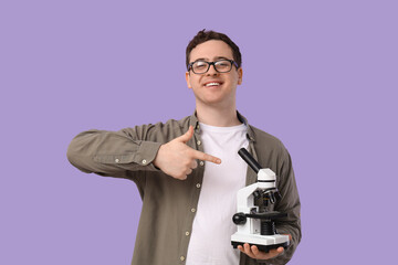 Handsome young man pointing at microscope on lilac background