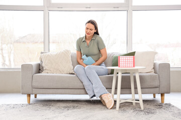 Young woman with hot water bottle and menstrual calendar suffering from cramps on sofa at home