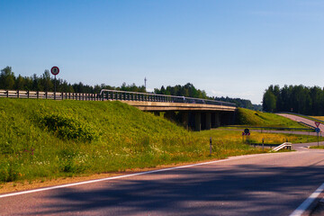 A concrete overpass stretches across a green landscape under clear skies, with traffic signs...