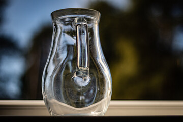 Clear glass pitcher placed on a windowsill, captured with soft natural light and blurred outdoor...