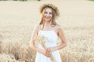 Portrait of a beautiful young woman in white dress and wreath of wheat ears in the field.