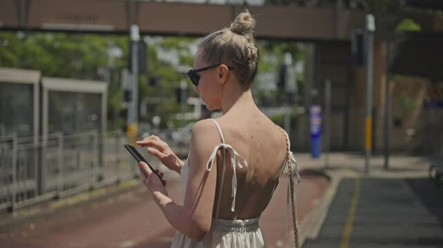A young attractive woman in a light backless dress stands at a city bus stop, using her smartphone for communication and browsing. Urban lifestyle scene with modern technology