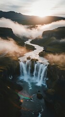 Fototapeta premium Spectacular Waterfall Cascading into a River Surrounded by Misty Mountains at Sunrise