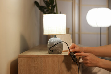 Woman turning off glowing lamp on table in bedroom at evening, closeup