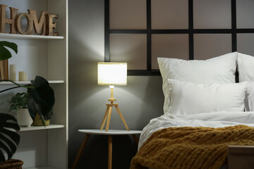 Interior of stylish bedroom with bed and glowing lamp on bedside table at evening