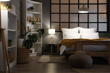 Interior of stylish bedroom with cozy bed, shelving units and glowing lamps at evening