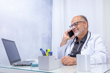 Smiling doctor in white coat sitting at a desk talking on the phone and looking at the computer