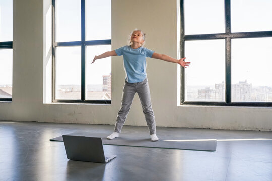 A person stretches joyfully in a bright room while following an online yoga class on a laptop