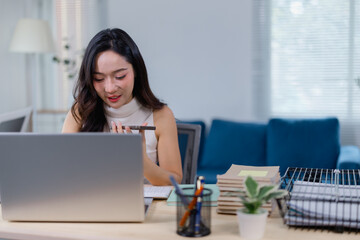 Young businesswoman using voice command technology at her desk, efficiently multitasking with a laptop, smartphone, and documents in a modern office environment