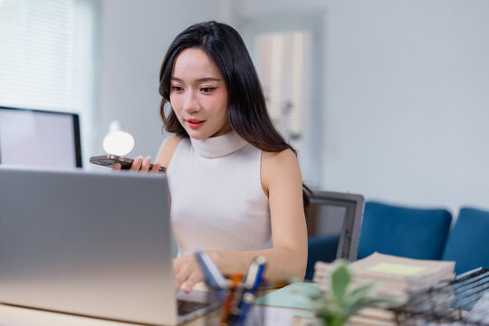 Young professional woman multitasking in an office setting, using voice command on her smartphone while working on a laptop. The environment is modern and organized, suggesting productivity