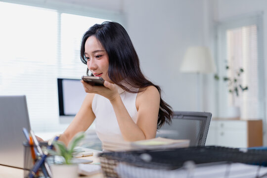 Professional woman multitasking at her office desk, using voice command on a smartphone while working on a laptop. Bright and modern workspace with office supplies and natural light - Powered by Adobe