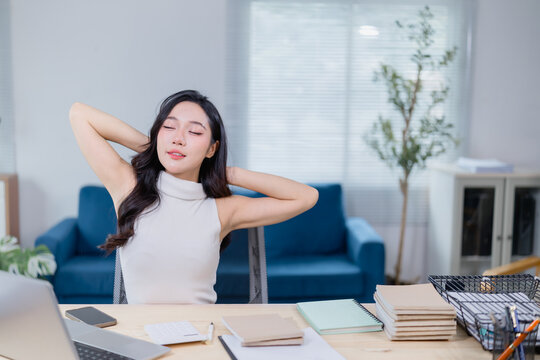 Young woman taking a break at her desk in a modern office setting. She leans back with eyes closed, surrounded by notebooks, a laptop, and office supplies, enjoying a moment of relaxation