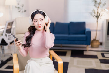 Young woman in a modern living room listening to music on headphones while holding a smartphone. She is seated comfortably in a stylish chair, enjoying a moment of relaxation