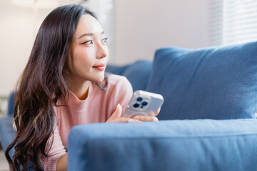 Young woman with long hair is relaxing on a blue sofa, holding a smartphone. She gazes thoughtfully...