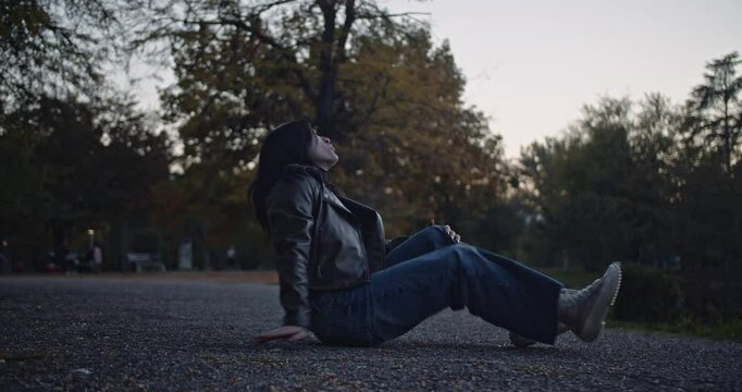 A young woman experiences excruciating pain after an unexpected fall in a park during the evening. She struggles to cope with the sudden injury, screaming and clutching her leg in distress.