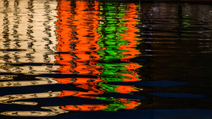 A close-up of a dark water surface showcases vibrant, rippling reflections of white, orange, and green lights, creating an abstract and mesmerizing pattern. This image is ideal for backgrounds or arti