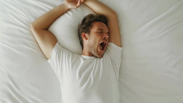 Sleepy male yawning, stretching in white bedroom setting, waking up with tired expression