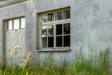 Broken vintage wooden window with shattered glass and abandoned door on weathered concrete wall of old rural building overgrown with tall summer grass and wildflowers in Latvia countryside