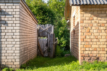 Old wooden gate between two brick buildings in a rural Latvian village, surrounded by lush green vegetation and summer sunlight, symbolizing passage, decay, and hidden stories