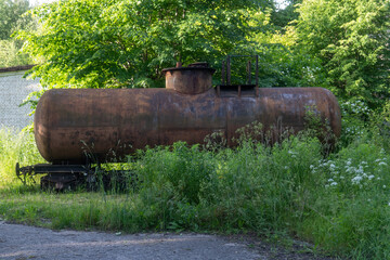 Rusty old fuel tank carriage overgrown by wild vegetation in abandoned area with green trees in...