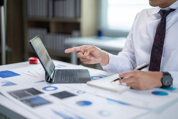 A man is pointing at a laptop screen while sitting at a desk with a notebook