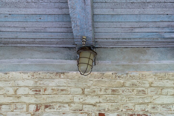 Close-up of old vintage industrial outdoor ceiling lamp with protective metal cage, mounted on weathered blue-painted wooden beams and brick wall of an abandoned building