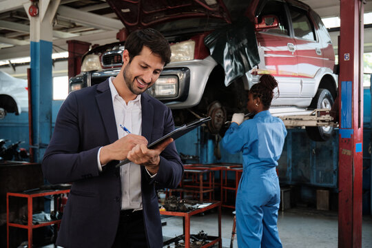 Cheerful Hispanic male mechanic manager with checklist documents for service insurance business, Black worker repair behind, car fix and claim warranty, maintenance at auto garage station industry.