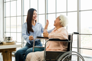 Obraz premium Young caregiver interacts warmly with a happy senior woman in a wheelchair near large windows, representing supportive elderly care, companionship, and assisted living in a healthcare environment.
