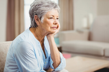 Senior woman sitting on sofa supporting cheek, gazing at coffee table at home, copy space