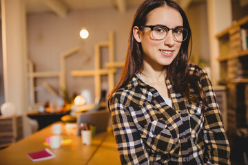 Woman standing behind wooden worktable in warm home office with mugs, notebooks, pens and laptop