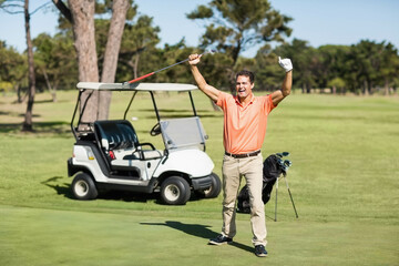 Golfer raising club in celebration on putting green near golf cart and bag under clear sky