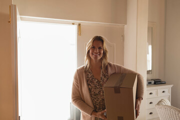 Woman standing near bright open doorway in entryway, holding moving box by dresser mirror, smiling