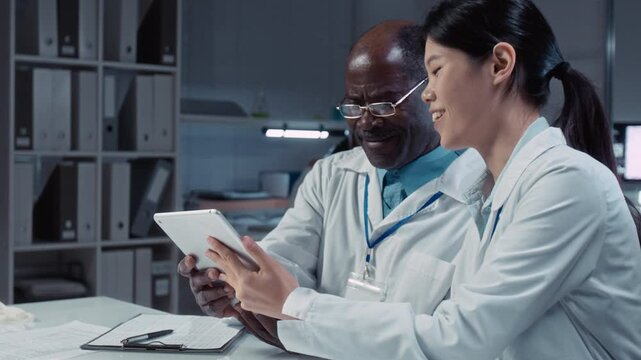 Medium shot of Chinese female student typing on tablet in laboratory, performing biochemistry research, showing result to mature African American male professor, correcting mistake and giving advice