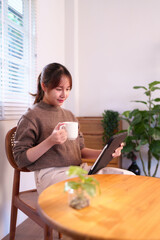 Relaxed Asian woman sips coffee and browses on tablet in her sunlit smart home integrating...