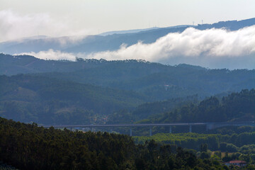 A misty mountainous landscape with layered clouds, dense forests, and a bridge
