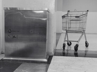 Black and white photo of a commercial space, featuring a shopping basket and a stainless steel trash can.