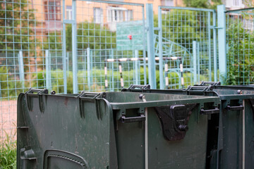 Row of green plastic trash containers positioned near a children’s playground, set against an old...