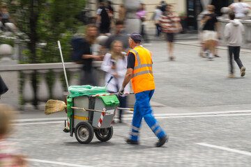 Male street cleaner wearing an orange safety vest and blue pants pushes a cleaning cart with tools...