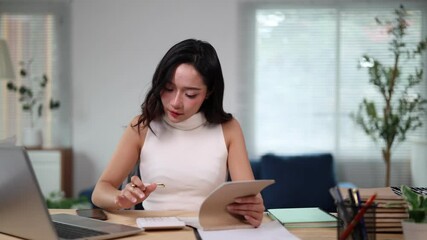 Professional businesswoman working remotely, analyzing data on digital tablet while taking detailed notes, demonstrating intense focus and concentration in modern home office setting - Powered by Adobe
