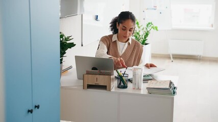 Female teacher flipping through pages in notebook and writing while sitting at desk and looking at laptop
