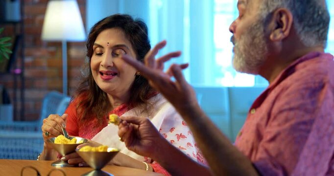 Senior couple eating ice cream at home, Indian Asian retired husband and wife enjoying sweet dessert treat together sitting on sofa or at round table, cheerful bonding moment with cone or bowl