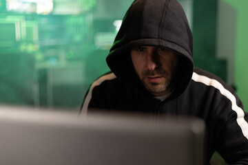 Hacker wearing a black hoodie is breaking into a secure network in a dark room with green code...