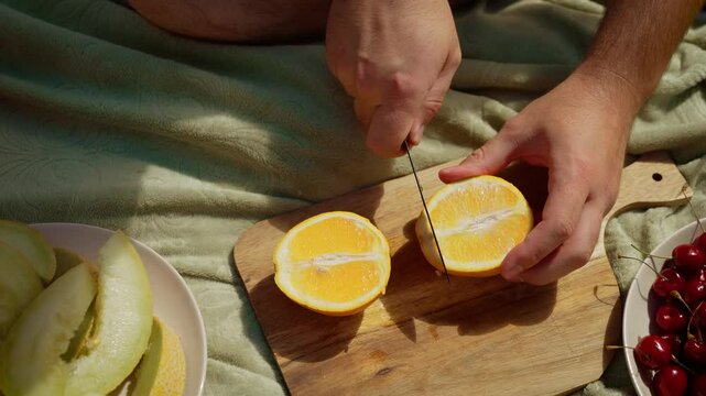 Man slicing orange on wooden cutting board, preparing fresh citrus for snacks. Male hands carefully dividing juicy orange into halves. Outdoor picnic setting with man cutting fruit for family