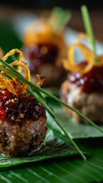Close up of gourmet meatball appetizers topped with red berry glaze, crispy fried onions, and chives, resting on vibrant green banana leaves.